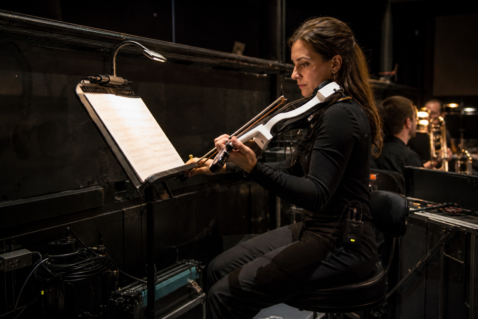 Седящ electric violin изпълнител чете от осветена стойка за ноти в theater-pit setup с организирани кабели и wireless pack наблизо