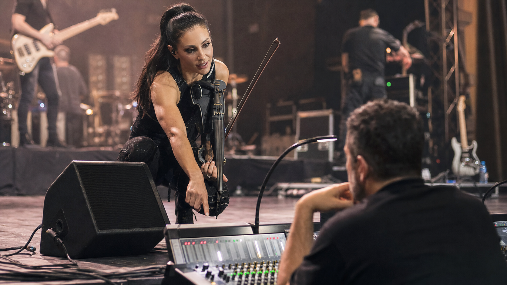 Electric violin изпълнител по време на full-band soundcheck сочи към floor monitor wedge, докато обсъжда нивата с front-of-house инженер